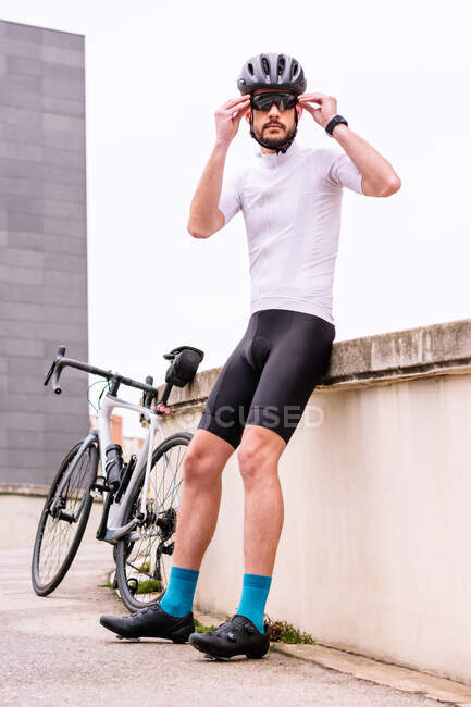 Бородатий чоловічий велосипедист у сучасних сонцезахисних окулярах та спортивному одязі проти міської будівлі під білим небом вдень — стокове фото