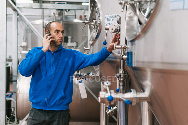 Vue latérale de l'entrepreneur masculin parlant sur un téléphone portable contre des navires en acier inoxydable sur le sol mouillé dans une usine de bière — Photo de stock
