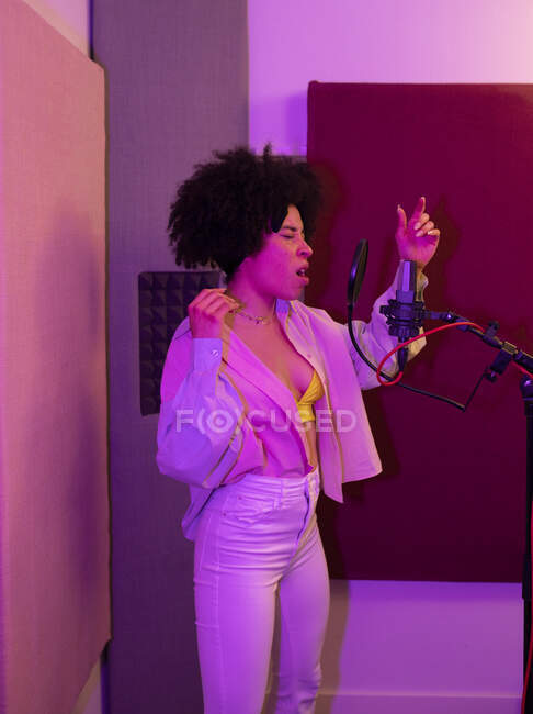 Cantora negra cantando música contra microfone com filtro pop enquanto está de pé com a mão no quadril e olhos fechados no estúdio de som — Fotografia de Stock