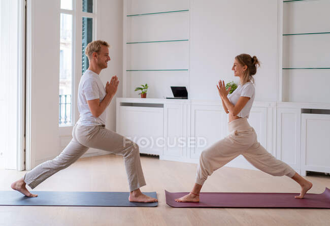 Seitenansicht eines zufriedenen Paares in Aktivkleidung, das auf Matten in Anjaneyasana steht, mit namaste Geste, während es morgens Yoga macht und sich gegenseitig ansieht — Stockfoto