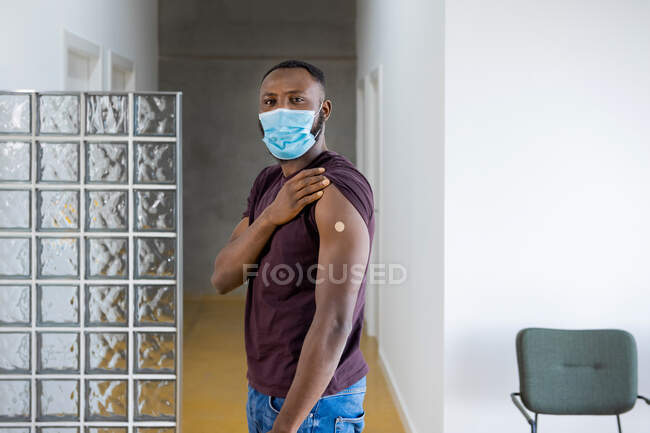 Hombre afroamericano con máscara protectora mirando a la cámara con orgullo mostrando yeso adhesivo vendaje en el brazo después de recibir la vacuna covid 19 de pie en una clínica durante el brote de coronavirus - foto de stock