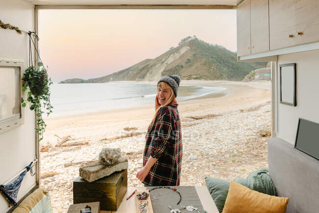 Vista lateral de la mujer viajera positiva artesana de pie cerca de camión con accesorios hechos a mano y mirando a la cámara en el fondo del mar y las montañas - foto de stock
