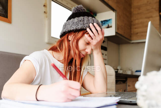 Side view of tired female traveler sitting at table in truck and taking notes while working on project remotely and leaning on hand — Stock Photo