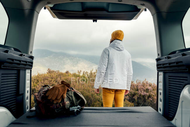 Back view of male camper in outerwear standing near van and admiring scenic view of highlands — Stock Photo