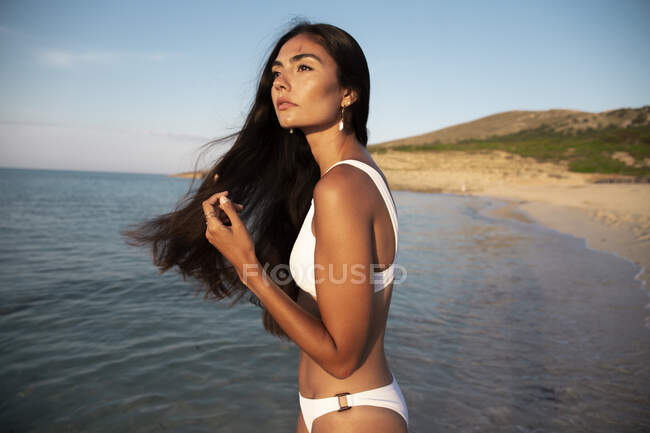 Vista lateral da jovem fêmea em roupa de banho branca e brinco olhando para o mar ondulado com montagem — Fotografia de Stock