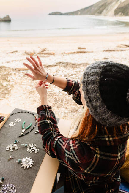 High angle of talented traveling female artisan trying on handmade bracelet while sitting at table in truck on seashore during trip — Stock Photo
