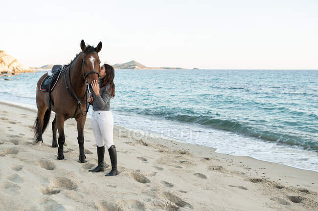 Young female kissing muzzle of chestnut stallion in bridle against wavy ocean — Stock Photo