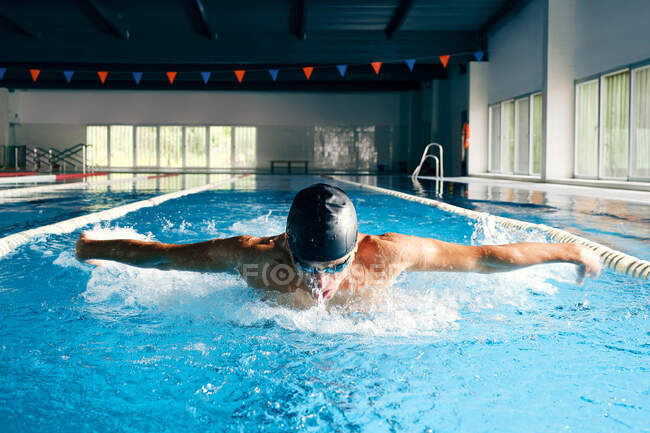 Starker männlicher Schwimmer mit Badekappe führt Schmetterlingsschlag beim Training im Schwimmbad mit blauem Wasser aus — Stockfoto