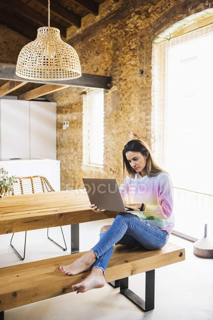 Barefoot female distance employee typing on netbook while sitting with crossed legs on wooden bench at home - foto de stock