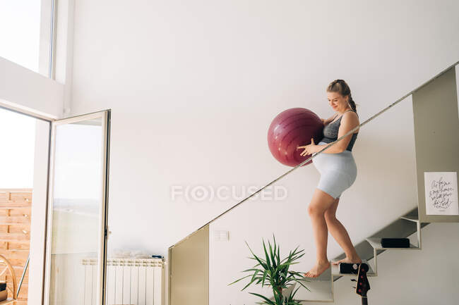 Side view of young smiling expectant female in sports clothes with yoga ball strolling on staircase in house — Stock Photo