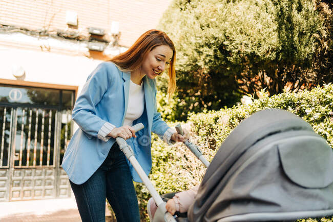Cheerful smiling mother wearing casual outfit talking to baby resting in stroller while standing on sunny street — Stock Photo