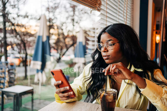 Elegante mujer afroamericana sentada a la mesa en la cafetería con refrescante bebida gaseosa y navegando por las redes sociales en el teléfono móvil - foto de stock