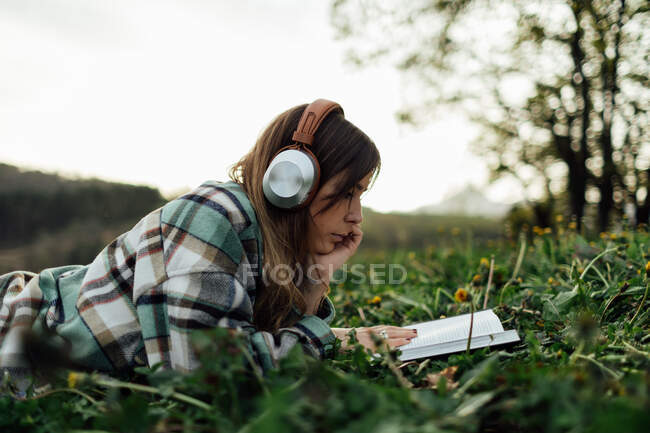 Side view of young attentive female in modern headset reading textbook while lying on meadow in summer — Foto stock