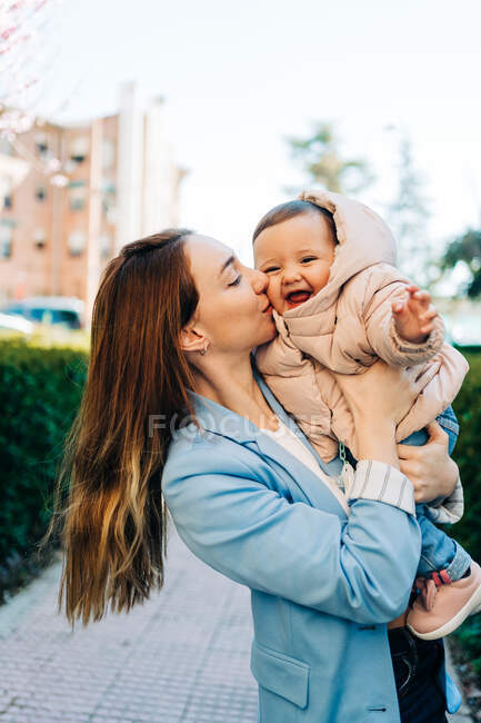Alegre joven madre en ropa casual que lleva y besa suavemente adorable bebé alegre mientras está de pie en la acera de la ciudad en el soleado día de primavera - foto de stock