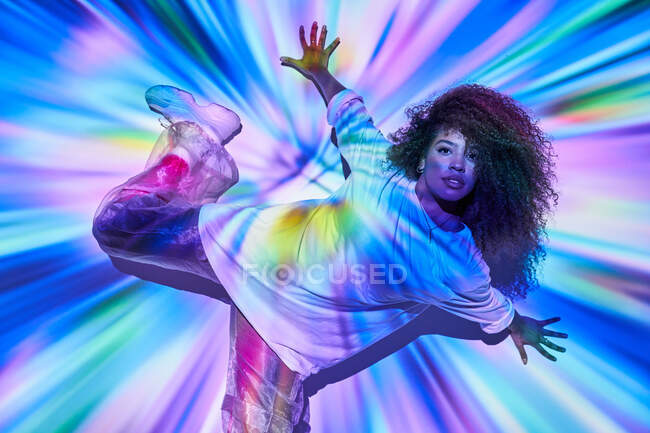 Энергичная афроамериканская танцовщица в неформальной одежде, танцующая на полу и смотрящая на камеру в неоновом свете в танцевальной студии — стоковое фото