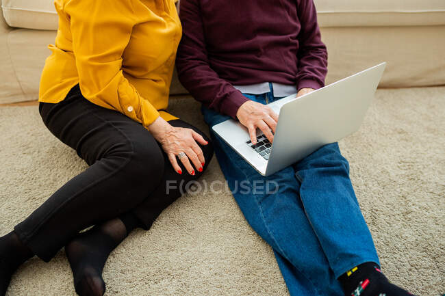 Crop anonyme couple d'âge mûr assis sur le sol à la maison et la navigation netbook ensemble — Photo de stock