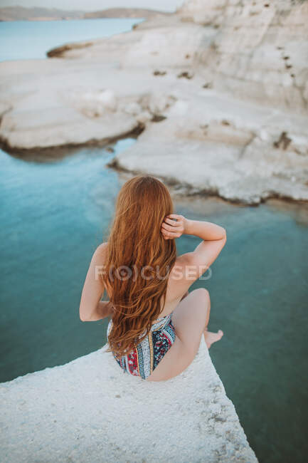 Back view unrecognizable fit female in stylish swimwear sitting on rocky cliff edge and admiring picturesque sea and shores views of Sarakiniko Milos while touching long hair — Stock Photo