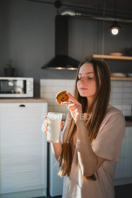 Joyeux jeune femme avec biscuit à l'avoine savoureux avec pépites de chocolat pour le petit déjeuner sur la table dans la cuisine — Photo de stock