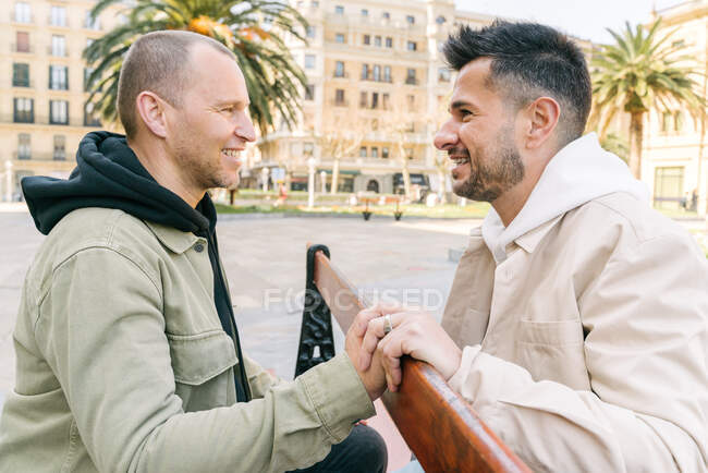 Side view of joyful young multiracial gay couple resting on wooden bench and looking at each other on city square on sunny day — Foto stock