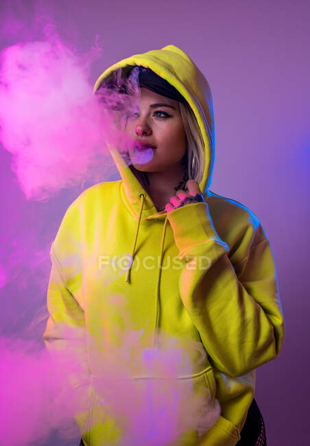 Mulher confiante hipster com capuz fumando e cigarro no estúdio em fundo rosa e olhando para longe — Fotografia de Stock