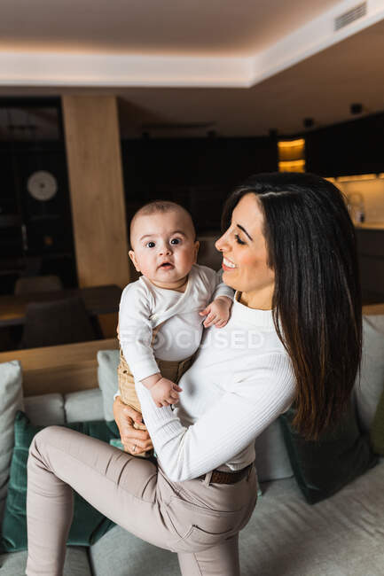 Вид сбоку на восхищенную мать, держащую восхитительно улыбающегося ребенка и веселящуюся вместе дома — стоковое фото