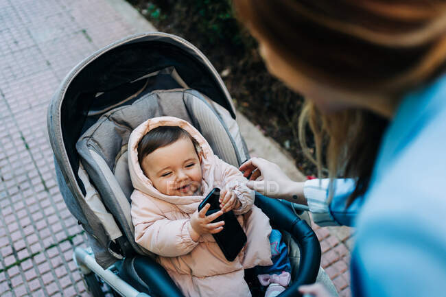 Сверху милый смешной ребенок со смартфоном в теплой одежде сидит в коляске и смотрит на размытую маму на весенней улице — стоковое фото
