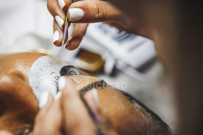 High angle of crop unrecognizable cosmetologist with tweezers applying fake eyelashes for extension on eye of ethnic client in salon - foto de stock