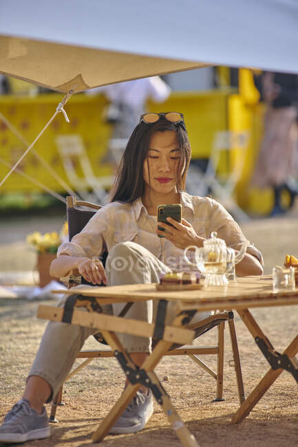 Menina asiática bonita que navega no telefone móvel ao ter um tempo relaxante sentado à mesa na área de acampamento — Fotografia de Stock