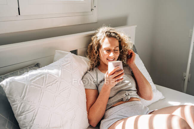 Happy young female in casual domestic clothes enjoying hot drink and having pleasant conversation via mobile phone while lying on comfy bed in light bedroom in morning — Photo de stock