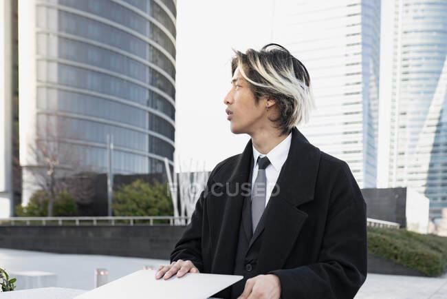 Jovem bem vestido executivo masculino étnico com acabamento de cabelo tingido trabalhando em netbook contra edifícios modernos da cidade olhando para longe — Fotografia de Stock