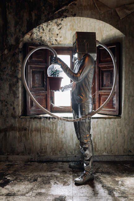 Вид збоку невпізнавана людина в срібному костюмі з дихаючим апаратом і шлангом, прикріпленим до горщика рослини, що стоїть в покинутій кімнаті — стокове фото