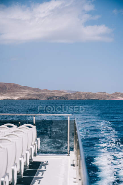 Пустые белые стулья на палубе круизного судна, плывущего в голубой морской воде с горой на берегу — стоковое фото