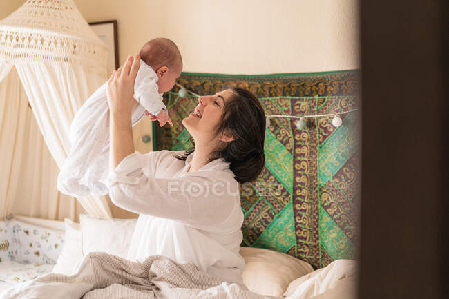 Smiling mom interacting with unrecognizable little child on crumpled bed at home in daylight — Stock Photo