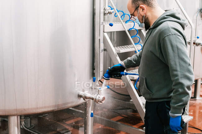 Side view of crop male worker in mask washing valve of metal tank at work in brewing company during pandemic — Stock Photo