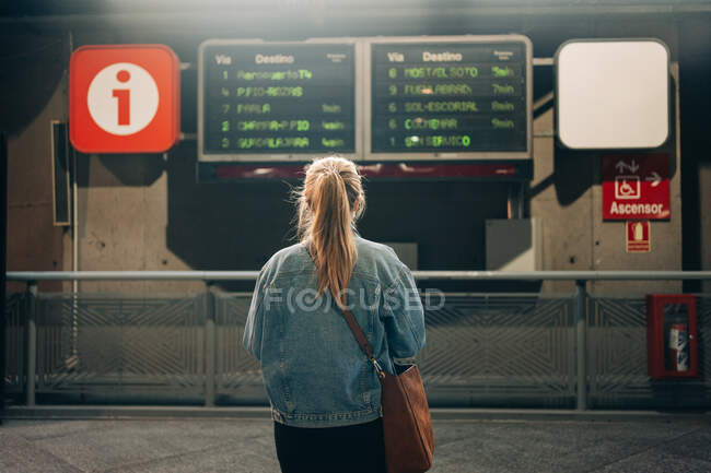 Зворотній вигляд жінки в джинсовій куртці і з сумкою, дивлячись на розклад поїздів на борту, що стоять на станції — стокове фото