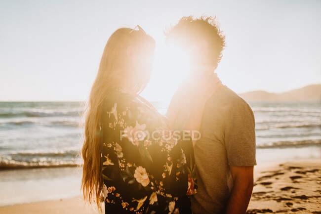 Mann umarmt junge attraktive Freundin in Badebekleidung, während er an einem sonnigen Tag in Fyriplaka Milos gemeinsam an der sandigen Meeresküste in der Nähe rauer Klippen chillt — Stockfoto