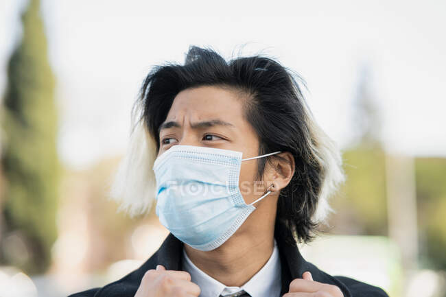 Joven ejecutivo masculino étnico con el pelo teñido con máscara desechable mirando hacia otro lado en la ciudad sobre fondo borroso durante el brote de coronavirus - foto de stock