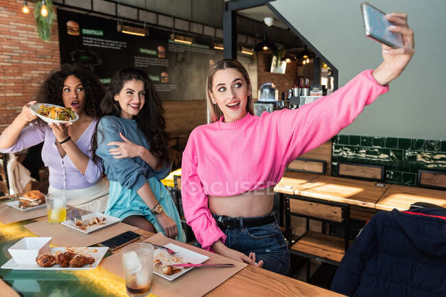 Feliz joven amigas con ropa casual tomando selfie en el teléfono móvil mientras almorzan juntas en un restaurante moderno - foto de stock