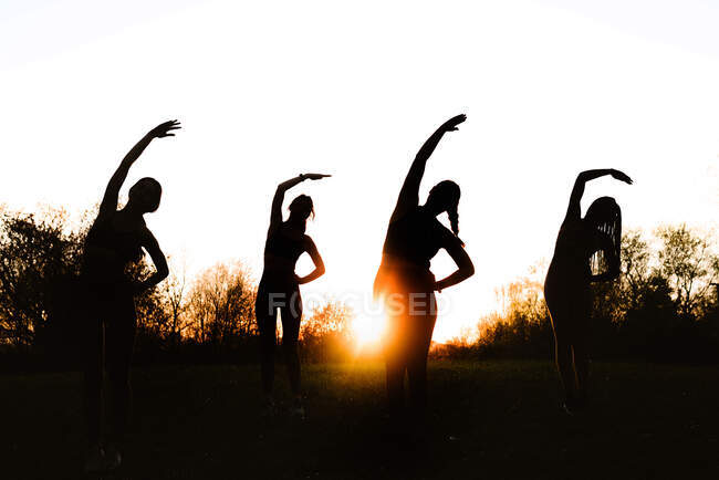 Baixo ângulo de silhuetas de atletas anônimos do sexo feminino fazendo exercício de curvatura lateral enquanto se alongam juntos no parque no fundo do céu por do sol — Fotografia de Stock