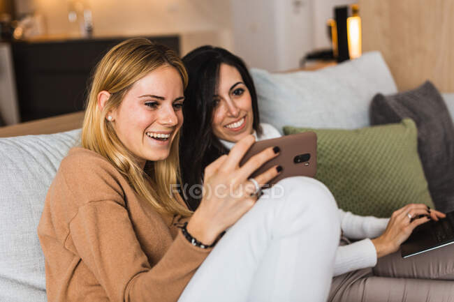 Couple of homosexual females sitting on sofa and watching funny video on mobile phone while laughing and having fun — Stock Photo