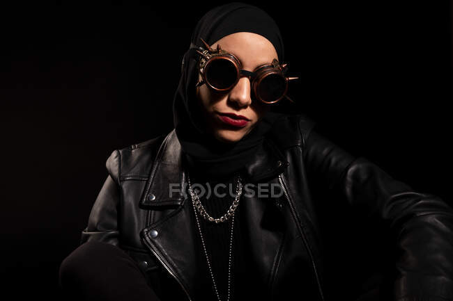 Уверенная молодая мусульманка в кожаном плаще и креативных солнцезащитных очках, сидящая на черном фоне в студии — стоковое фото