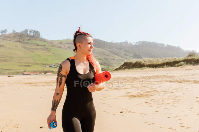 Улыбающаяся спортсменка с катающимся ковриком и бутылкой воды, прогуливаясь по песчаному морскому побережью, отворачивается — стоковое фото