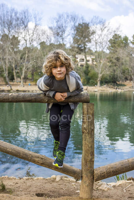Child in casual outfit on old wooden fence looking at camera against rippled water and trees — Photo de stock