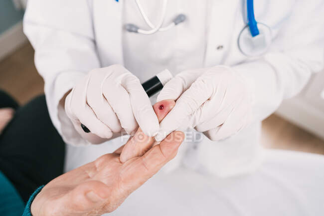 De arriba del cultivo el médico irreconocible en los guantes estériles tomando el análisis de sangre del paciente con la lanceta en el hospital - foto de stock