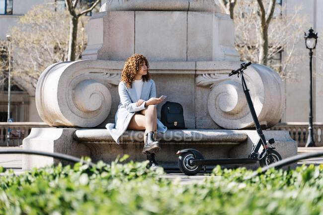 Full body young African American female in blue coat browsing on mobile phone while sitting on stone bench in spring park near scooter — Stock Photo