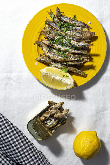 From above of fried and canned anchovies served on table with fresh lemons in restaurant in sunlight - foto de stock