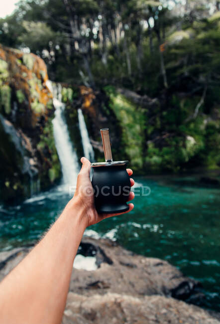 Mano de corte de macho con mate en taza de calabaza de pie sobre el fondo de la cascada en el bosque - foto de stock