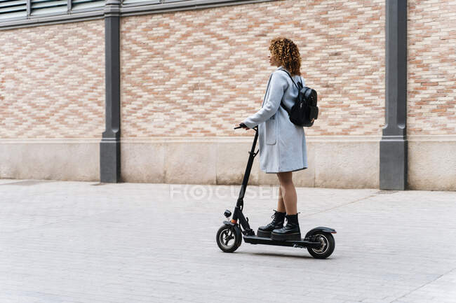 Full body side view of content African American young female in casual wear riding scooter on paved sidewalk and looking away with smile — Photo de stock