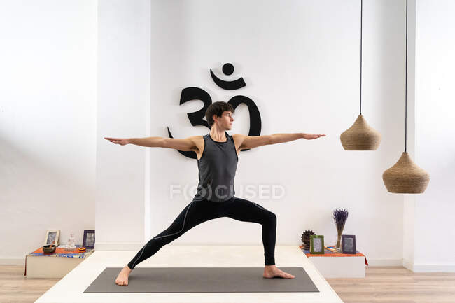 Anmutiger junger Mann steht im Virabhadrasana und praktiziert Yoga auf Matte im Studio — Stockfoto