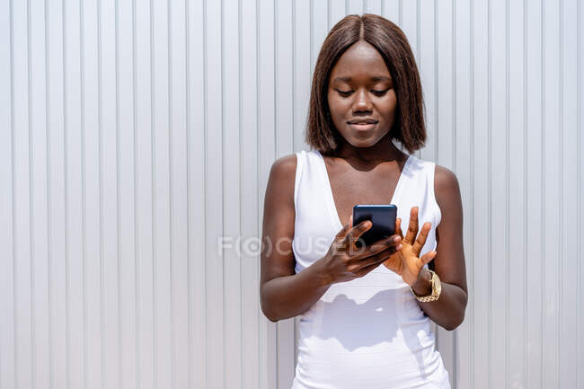 Positive stylish African American female in white dress browsing modern mobile phone while standing near building wall on sunny street — Stock Photo